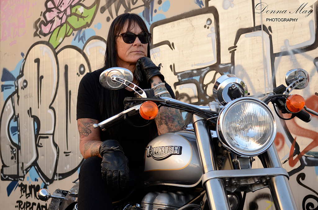 A photograph of a woman sitting on her motorbike in front of a graffiti wall