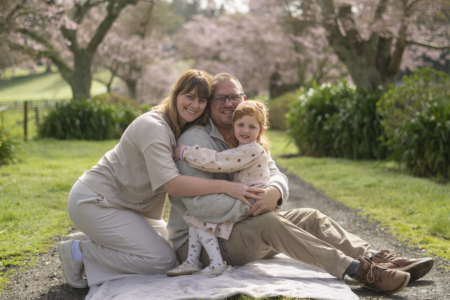 A photograph of a young family sat under cherry blossom trees. Taken by Te Awamutu, Waikato based photographer, Donna from Donna May Photography.