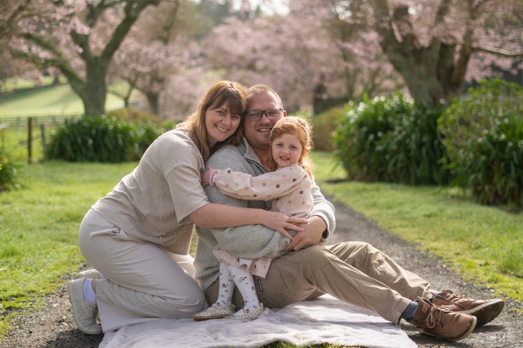 A photograph of a young family sat under cherry blossom trees. Taken by Te Awamutu, Waikato based photographer, Donna from Donna May Photography.