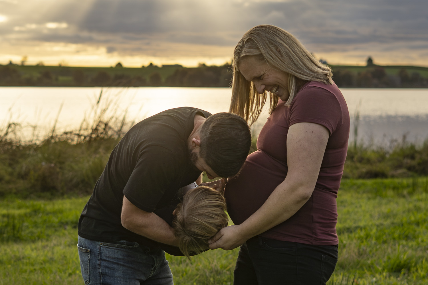 A family maternity photograph of an expectant mum with her husband and young son next to a lake in Te Awamutu. Taken at golden hour by Donna from Donna May Photography. A Waikato, Te Awamutu based lifestyle family photographer.