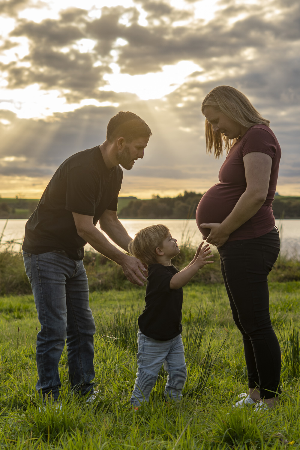 A family maternity photograph of an expectant mum with her husband and young son next to a lake in Te Awamutu. Taken at golden hour by Donna from Donna May Photography. A Waikato, Te Awamutu based lifestyle family photographer.