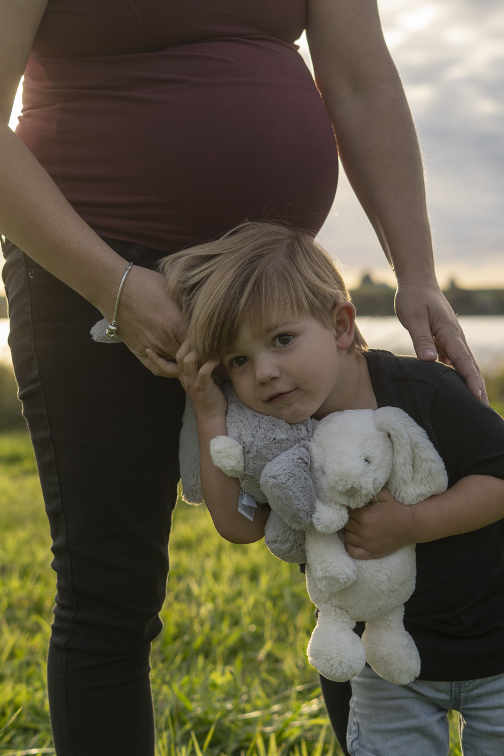 A family maternity photograph of an expectant mum holding her toddlers hand. Taken by a lake in Te Awamutu. Photo taken at golden hour by Donna from Donna May Photography. A Waikato, Te Awamutu based lifestyle family photographer. 