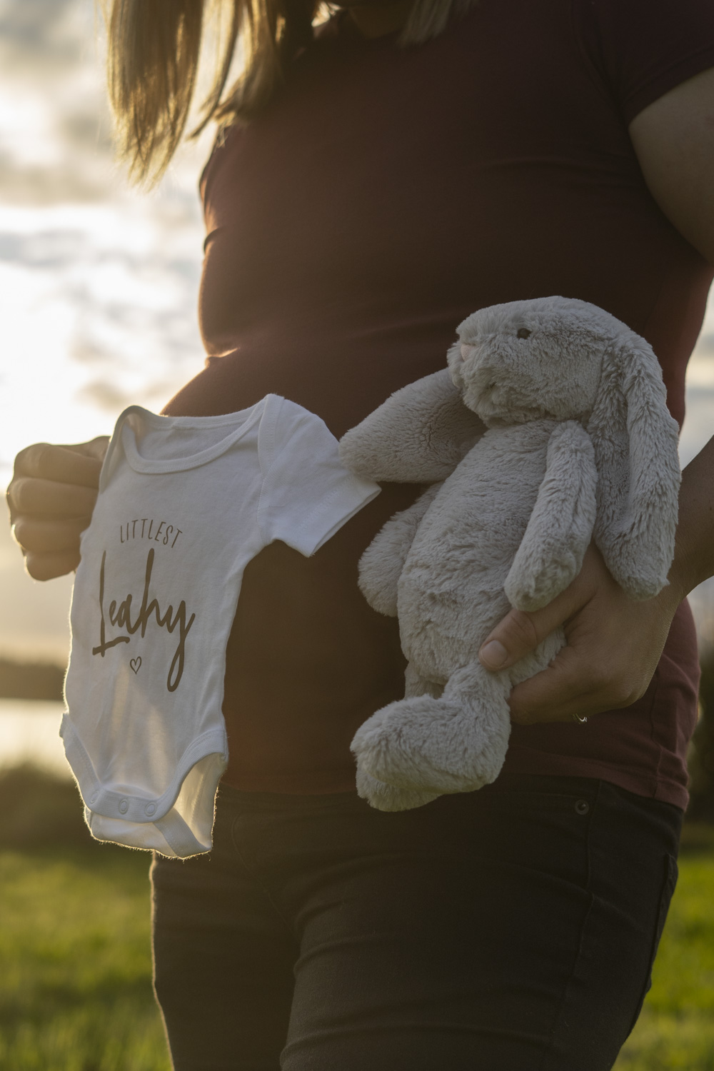 A maternity photo of an expectant mum holding a bunny and baby grow next to her bump. Taken at golden hour by Donna from Donna May Photography. A Waikato, Te Awamutu based lifestyle family photographer.