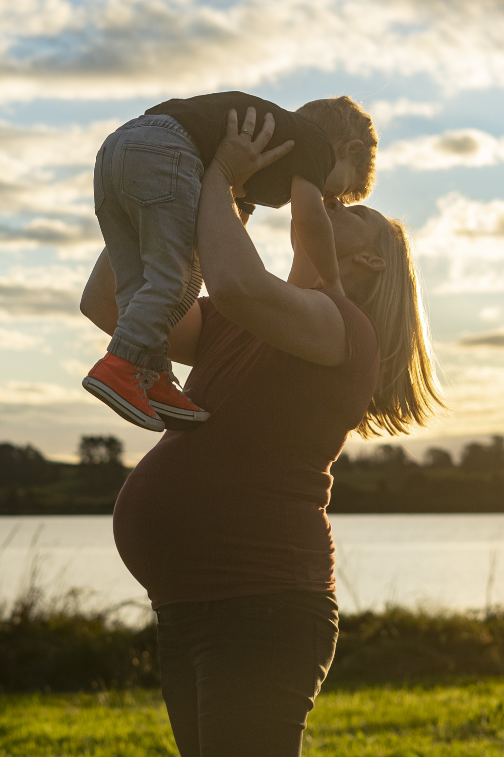 A family maternity photograph of an expectant mum holding her toddler in the air whilst giving him a kiss. Taken by a lake in Te Awamutu. Photo taken at golden hour by Donna from Donna May Photography. A Waikato, Te Awamutu based lifestyle family photographer. 