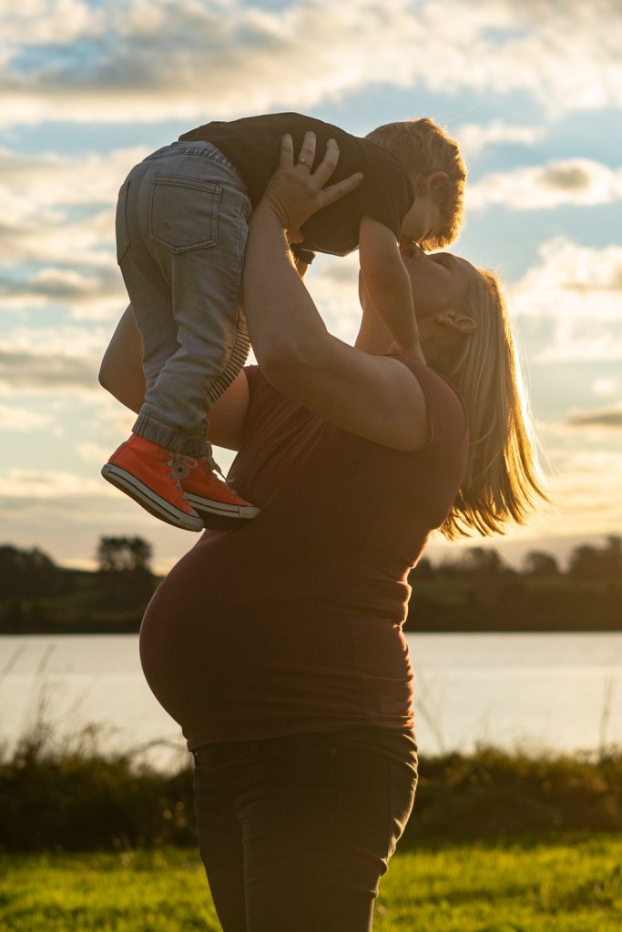 A family maternity photograph of an expectant mum holding her toddler in the air whilst giving him a kiss. Taken by a lake in Te Awamutu. Photo taken at golden hour by Donna from Donna May Photography. A Waikato, Te Awamutu based lifestyle family photographer.