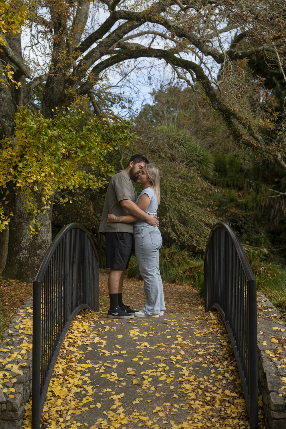A portrait photo of a couple sharing a kiss whilst standing on a park bridge in autumn. Taken by Te Awamutu, Waikato based photographer, Donna from Donna May Photographer